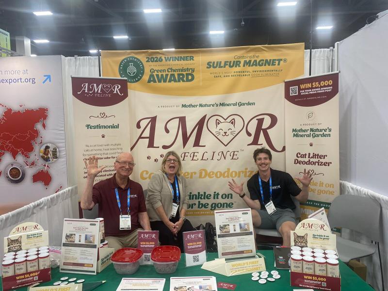 Three members of the AMOR FELINE team waving from the booth in front of the Green Chemistry Award banner
